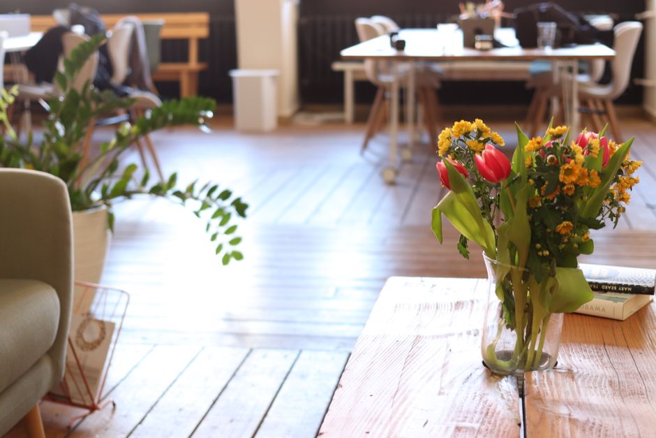 A glass vase filled with a fresh flower bouquet featuring pink tulips, yellow daisies, and green foliage, placed on a light wooden table inside a modern office or café setting. The arrangement is wel