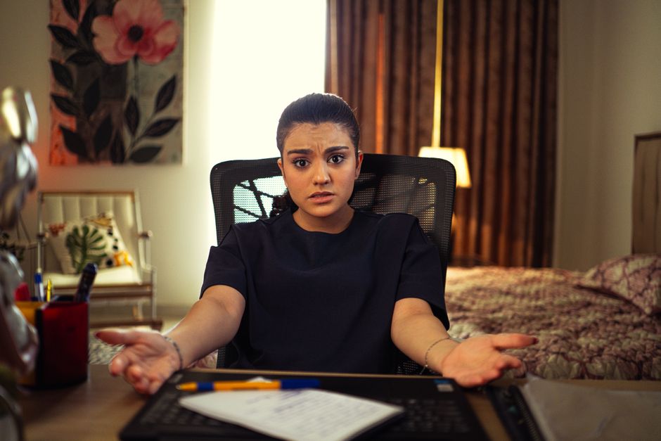 A young woman with dark hair tied back, sitting at a desk in an office room, showing a confused or concerned expression with her eyebrows furrowed and palms turned upward. Behind her, there is a bed w