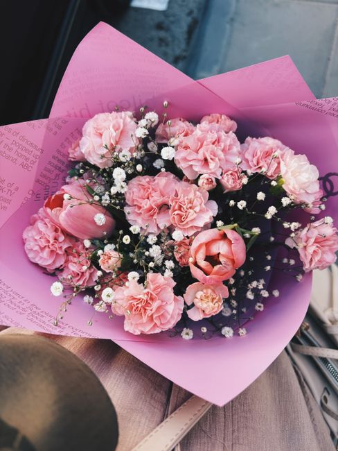 A vibrant bouquet of fresh pink flowers, including carnations, tulips, and smaller filler blooms like gypsophila, wrapped in pink tissue paper. The arrangement is held in hand, with the pink wrapping