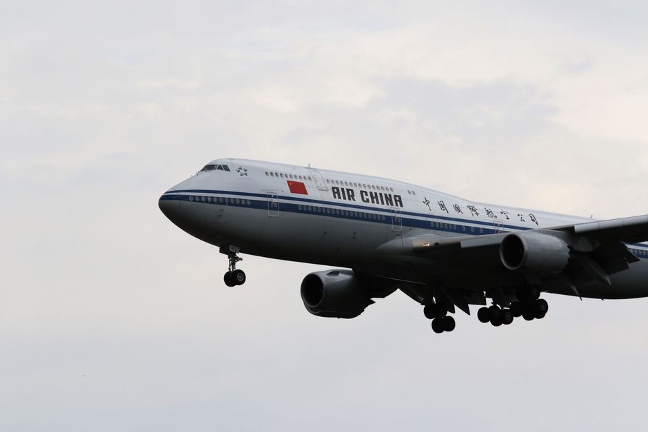 High-angle photograph of an Air China commercial airplane in flight against a cloudy sky, showing the aircraft's nose, cockpit windows, and part of the fuselage with the airline's logo and Chinese cha