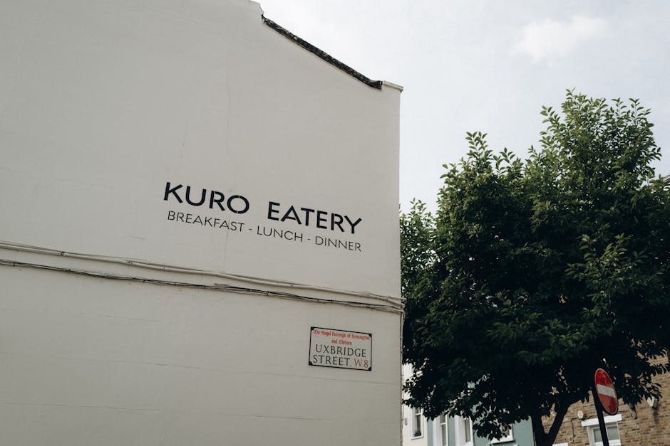 A close-up view of a white exterior wall with the name 'KURO EATERY' prominently displayed, indicating a restaurant offering breakfast, lunch, and dinner. Adjacent to the signage, a street sign reads